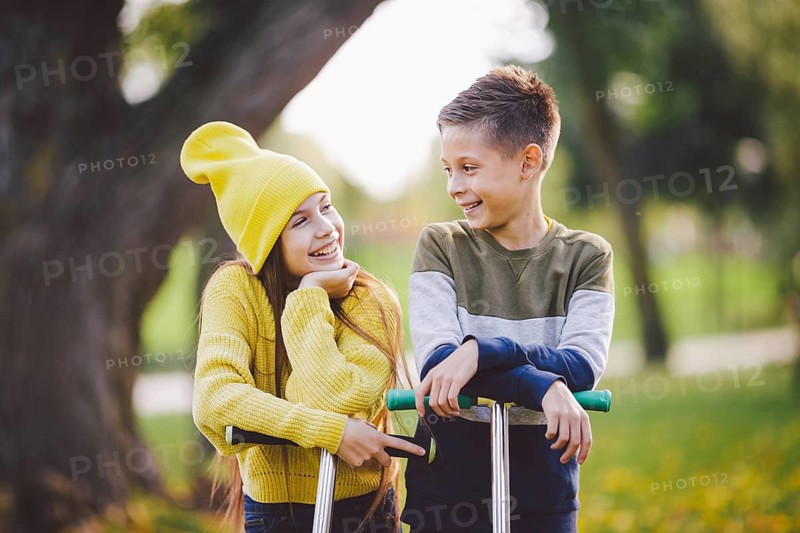 Twins 10 years old in bright clothes spend time actively together in autumn park riding kick scooters. Happy children. Eco transport. Kids posing in fall park. Brother and sister pre teens portrait