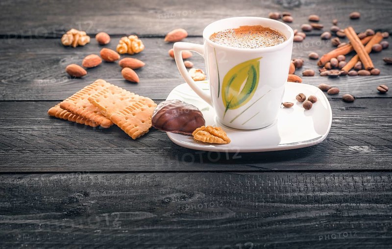 Cup of hot coffee on a plate, surrounded by tasty and healthy snacks, biscuits, almonds, walnuts and coffee beans, on an aged wooden table, in the morning light
