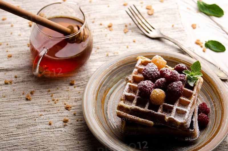 Belgian waffles served with raspberries and mint leaf dusted with powdered sugar on white wooden kitchen table with syrup aside. Selective focus