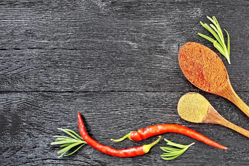 Frame of sprigs of fresh rosemary, pods of red hot pepper and two spoons with ground pepper on a wooden board background from above