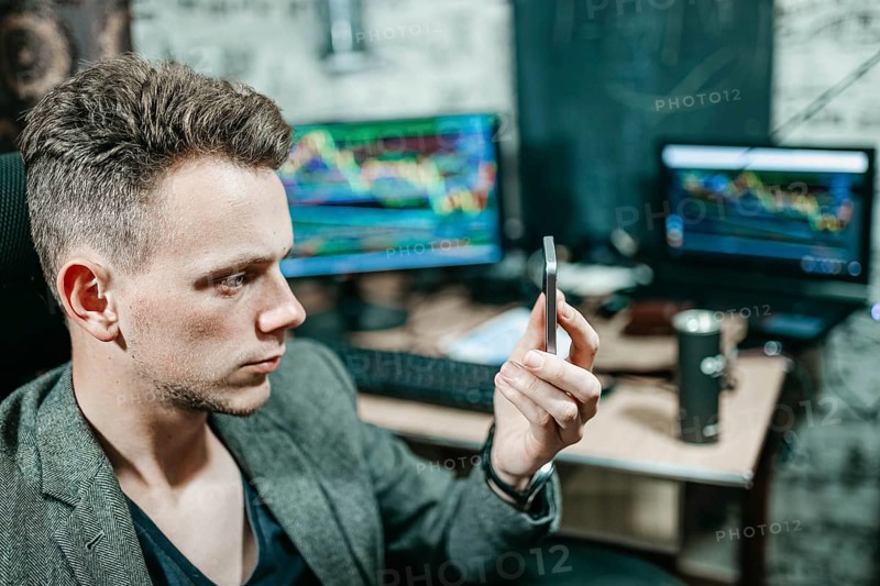 Man examining a smartphone in an office setting with trading monitors behind