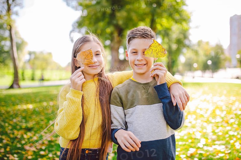 Happy Caucasian siblings enjoying in autumn day in park, sunny weather. Kids having fun holding fallen yellow leaves in nature. Funny twins in autumn park. Autumnal mood. Teenagers posing embracing