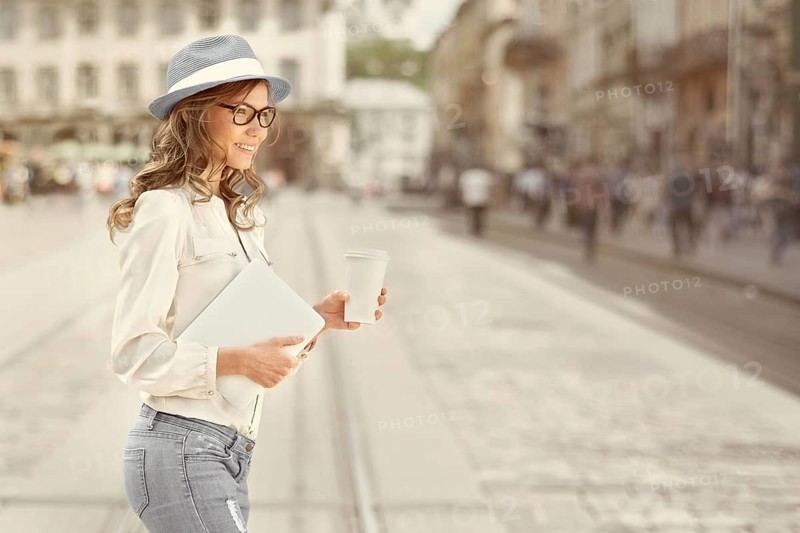 Beautiful young woman with a disposable coffee cup drinking coffee, holding a tablet in her hands and crossing the street against an urban background