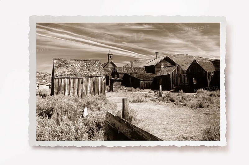 A dusty path between thorny undergrowth leads to wooden houses in the former gold mining town of Bodie. Photo in sepia tones with white jagged edges in the character of old photos, lying shadow. A dusty path between thorny shrubs leads to wretched wooden houses of the former gold mining town Bodie. Photo in sepia tone with white jagged edges