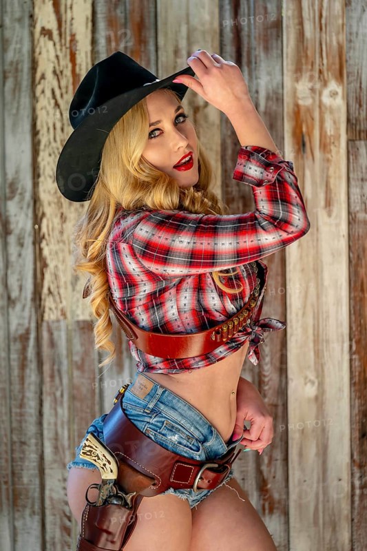 A confident, beautiful blonde woman in classic cowboy attire stands tall, posing with a gleaming pistol on her hip and a rifle resting across her shoulder. Her smile radiates energy and fearless charm, Henly, United States of America