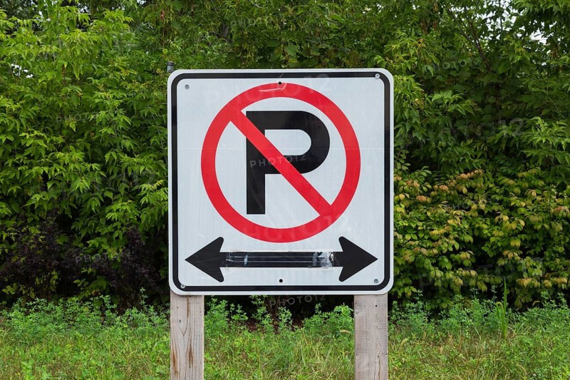 Large white, black and red No Parking with Left and Right direction arrows pictogram notice sign mounted on wooden posts in a nature park parking lot in summer, Quebec, Canada, North America