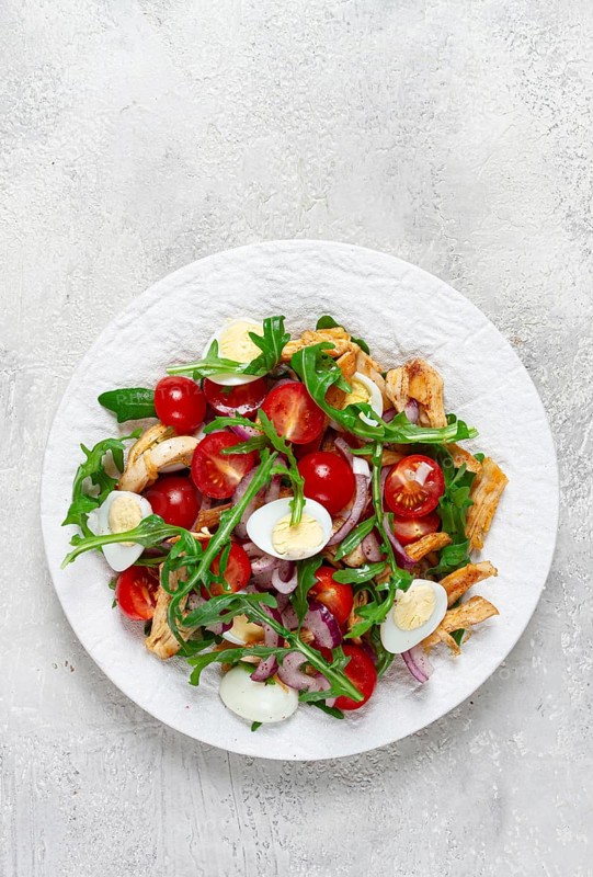 A warm salad with fried chicken fillet, arugula and cherry tomatoes, and boiled quail eggs, on a white plate, no people