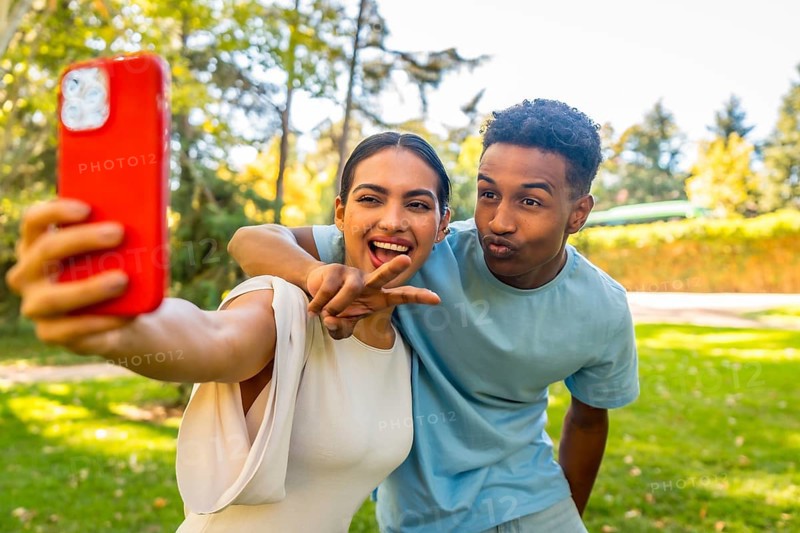 Multi-ethnic couple taking selfie gesturing victory and blowing a kiss in a park