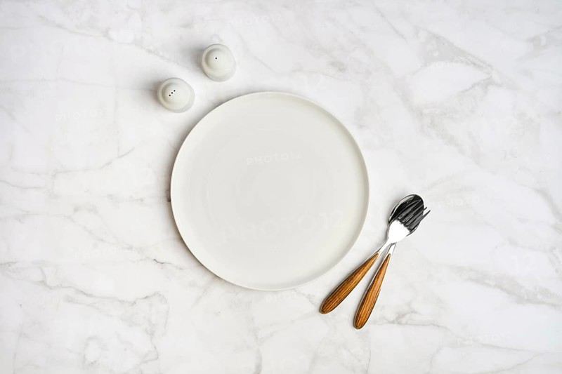 A simple dining arrangement showcases an empty white plate accompanied by a fork and knife with wooden handles, placed on a smooth marble surface, alongside salt and pepper shakers