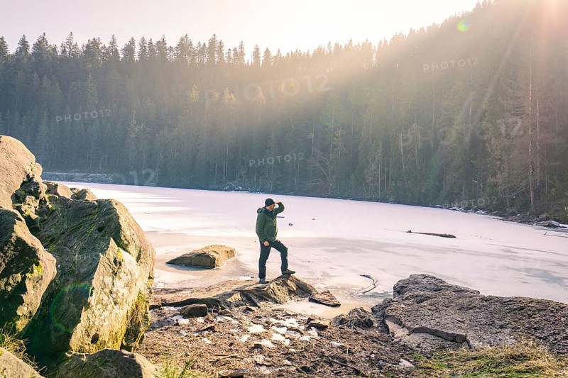 Person standing on the shore of a frozen lake surrounded by forest and sunbeams, Glaswaldsee, Bad Rippoldsau-Schapbach, district of Wolfach, Black Forest, Germany, Europe