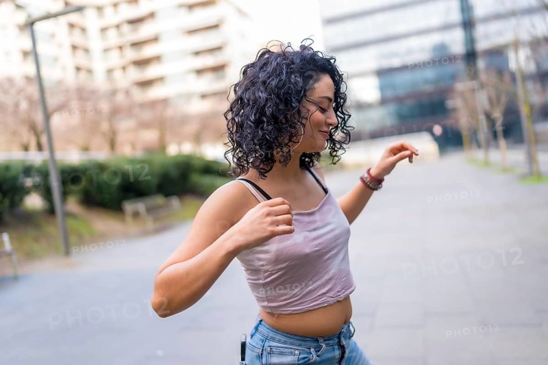 Curly haired hispanic woman joyfully dancing in an urban setting, embracing the sunny day with vibrant energy and carefree movement