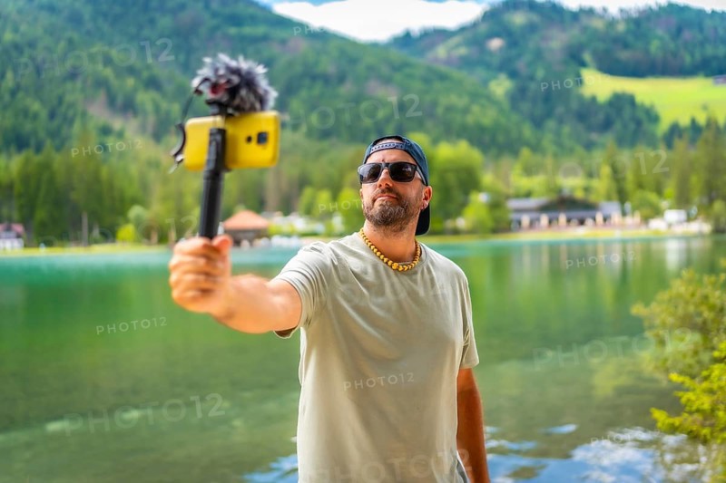 Content creator filming a video with a smartphone and external microphone at lake dobbiaco in the italian dolomites during summer