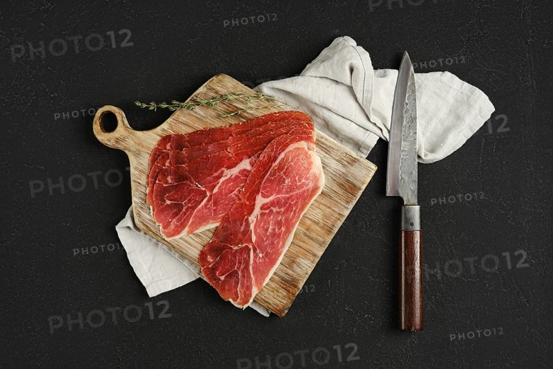 Overhead view of jamon iberico cured pork meat on a wooden cutting board, accompanied by a sharp knife. A soft cloth adds texture, while a sprig of thyme enhances the presentation, suggesting a culinary preparation.
