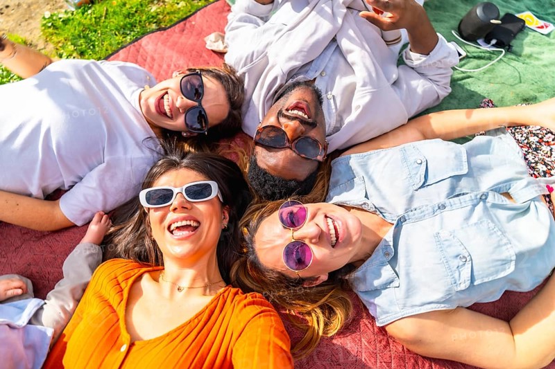 Four cheerful young adults lying together on blanket, enjoying sunny day picnic in park, listening to music and smiling