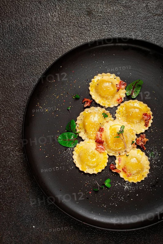 Fresh ravioli, with butter, spices and herbs, fried bacon, on a black plate, top view, dark background, no people