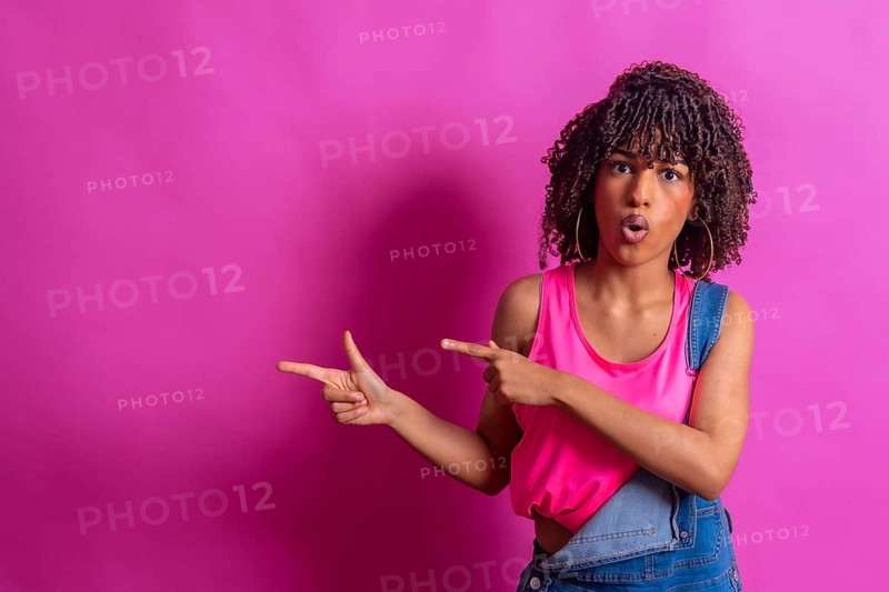 Surprised young woman with curly hair points to the side with a shocked expression, suggesting a promotional opportunity against a vibrant pink backdrop
