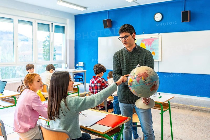Geography teacher engaging students by pointing out countries on a globe during an interactive lesson in a lively classroom setting