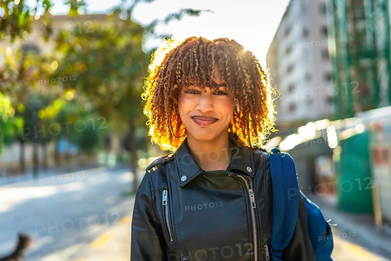 Confident young latin woman with curly hair and leather jacket smiling at camera standing in the street during sunset