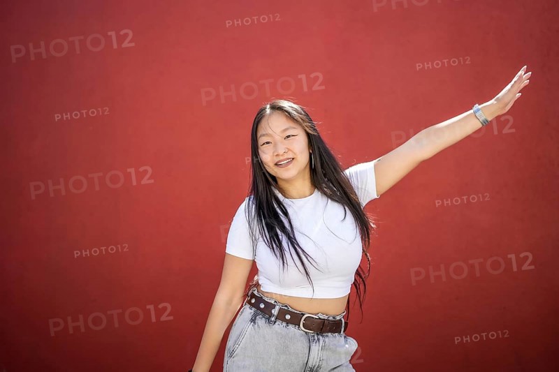 Frontal portrait of a happy beauty young asian woman raising arm dancing K-pop against a red urban wall