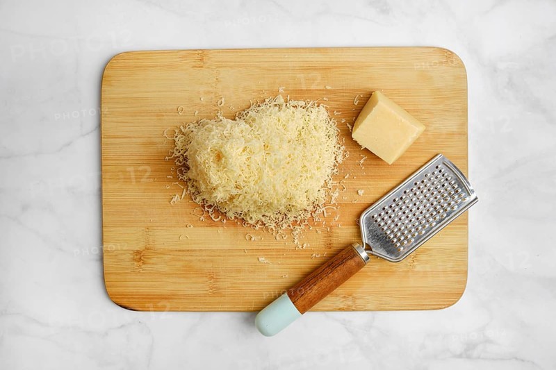 Shredded cheese on a wooden cutting board alongside a block of cheese and a grater. The items are arranged neatly in a well-lit kitchen, suggesting preparation for a delicious meal