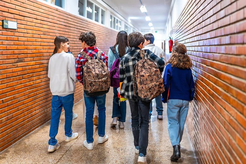 Rear view of diverse group of students walking down school hallway between classes, carrying backpacks and chatting
