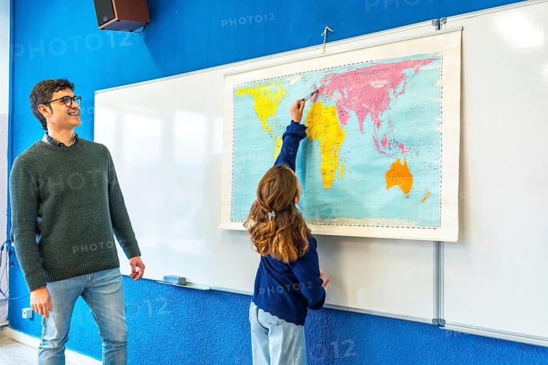 Smiling teacher watching elementary school student pointing at a world map during geography class