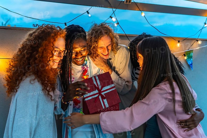 African man opening birthday present embraced by his multiracial friends during rooftop party