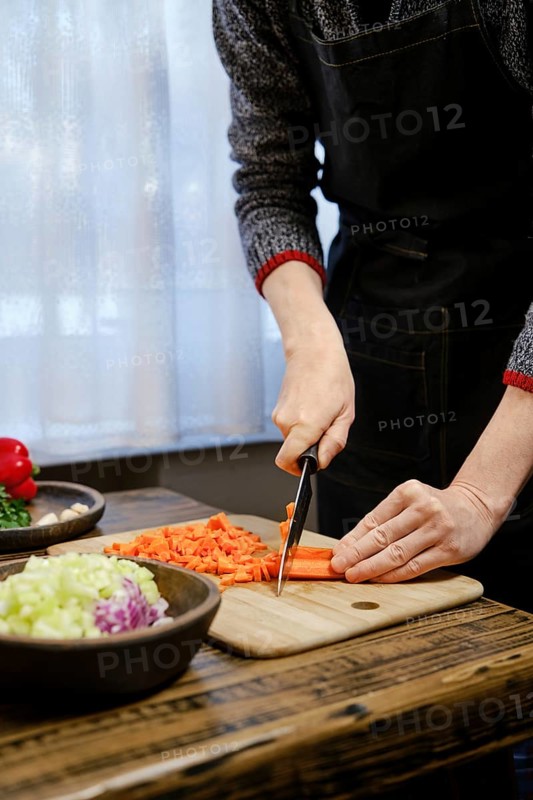 Unrecognizable man is chopping vibrant carrots on a wooden cutting board while surrounded by fresh vegetables like bell peppers and celery. Bright light filters through the window, creating a warm atmosphere.