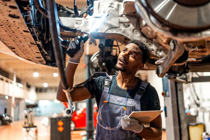 Young mechanic inspecting car chassis using flashlight and holding tablet in a modern workshop