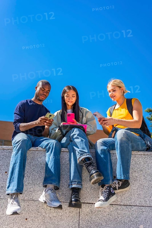 Vertical low angle view photo with copy space against blue sky of multi-ethnic students sitting on a wall outside the campus using mobile phones