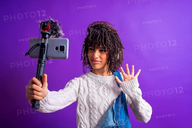 Young influencer waving while recording a video blog using a smartphone with external microphone on purple background
