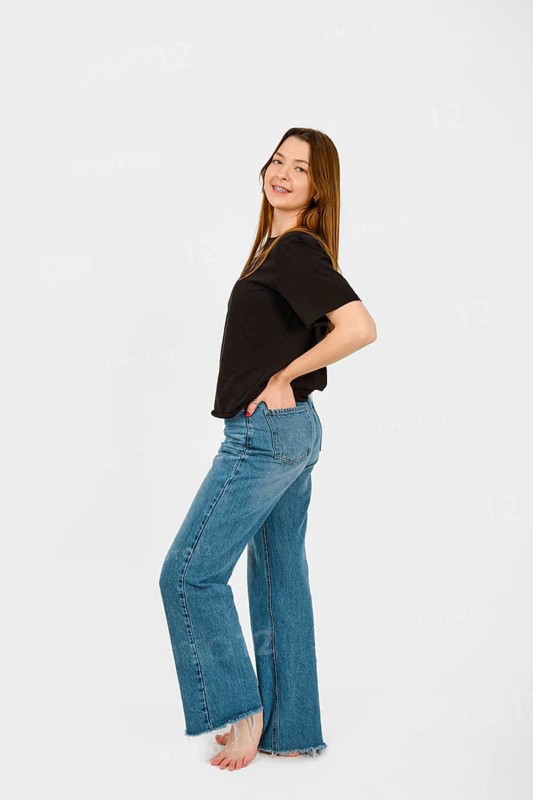 Barefoot woman stands with a playful posture, smiling at the camera while wearing a black top and wide-leg jeans. The simple white background emphasizes her casual, confident demeanor and style