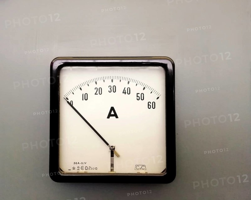 A large square industrial ammeter with an analogue dial with numbers with standard electrical symbols on a white dial on a grey background