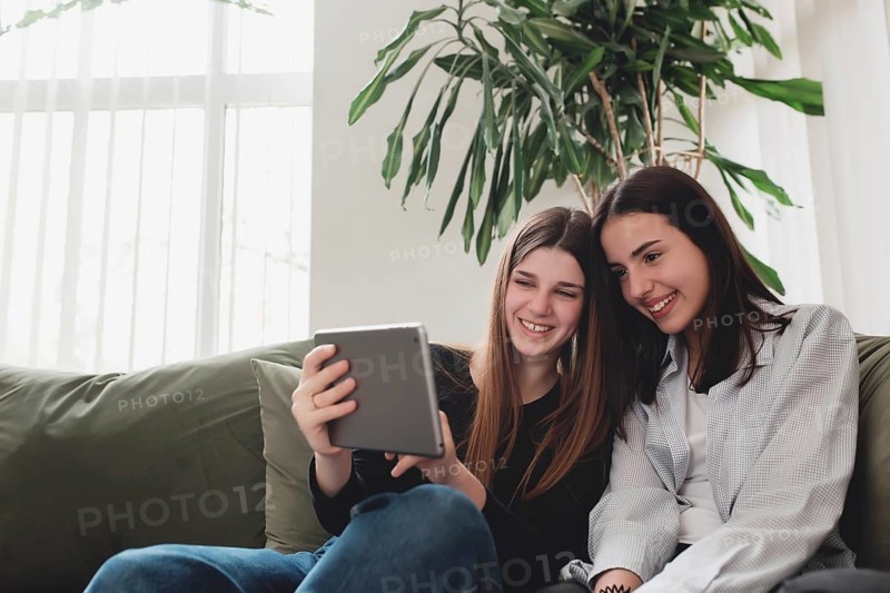 Two teenager sister girls are sitting at green sofa in living room and watching some funny video at tablet, smiling, laughing. Beautiful people, frendship and togetherness concept. Flowerpot on back