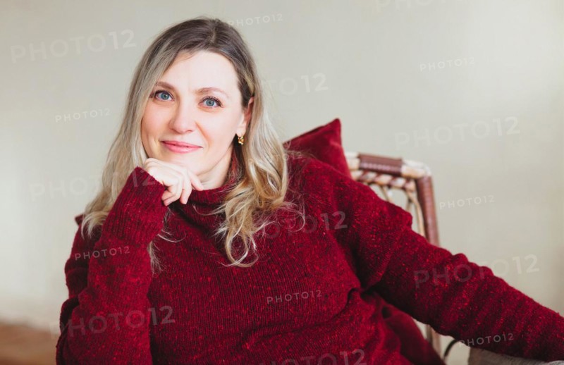 Attractive middle aged happy woman sitting and relaxing on comfortable chair at home on weekend, wearing colorful knitted warm red sweater, smiling at camera. People lifestyle lifestyle
