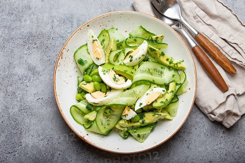 Healthy green avocado salad bowl with boiled eggs, sliced cucumbers, edamame beans, olive oil and herbs on ceramic plate top view on grey stone rustic table background, food photography, food photography