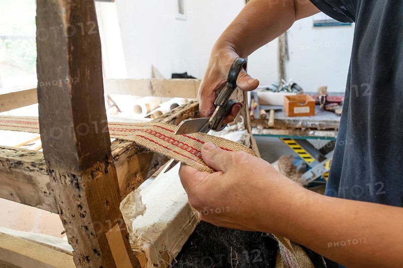 Upholsterer with traditional craft skills shortens a new strap on an old chair with scissors in his workshop, copy space, selected focus, narrow depth of field