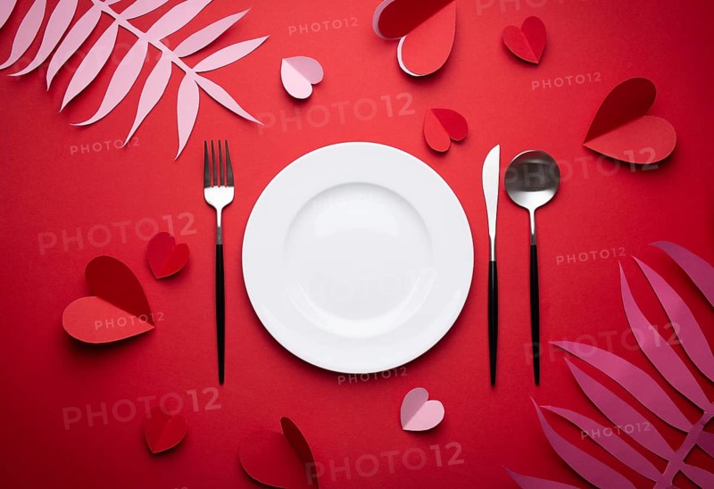 Romantic dinner at restaurant on Valentines day, paper art. White empty plate, spoon, fork, knife on red background with paper hearts and tropical leaves. Valentines day date concept craft origami