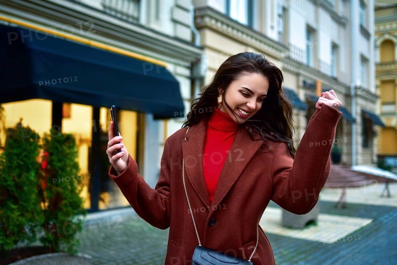 Beautiful Overjoyed young brunette woman with long hair in a red coat celebrating good news holding phone and clenching fist in the evening street