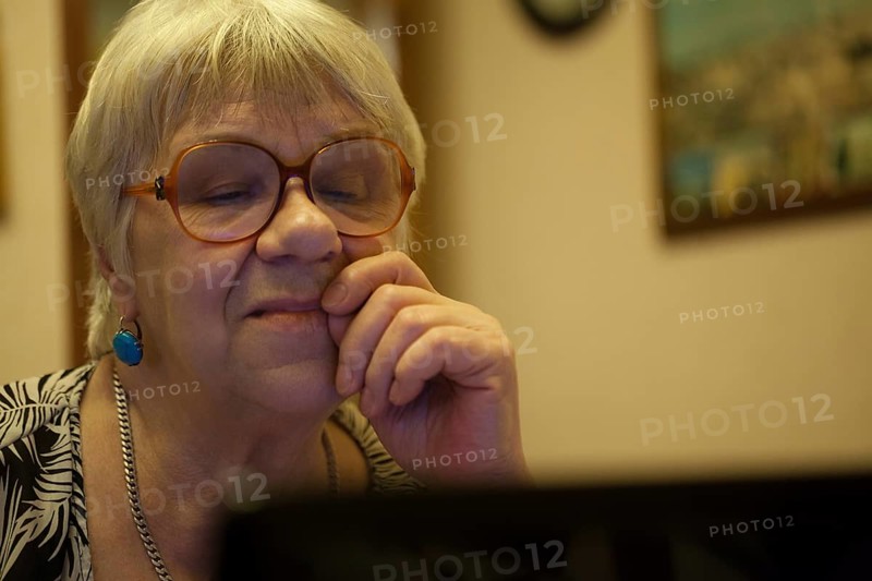 Thoughtful senior woman with blond hair wearing spectacles reading a computer screen with her hand to her face and an expression of concentration