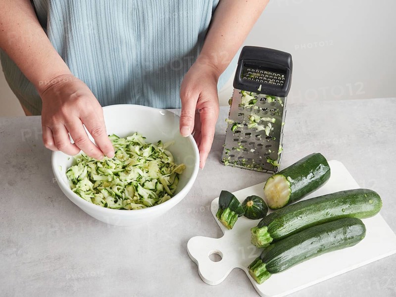 Woman with her hands in a bowl of grated zucchini, other fresh zucchini on wooden board and stainless steel hand grater on the countertop
