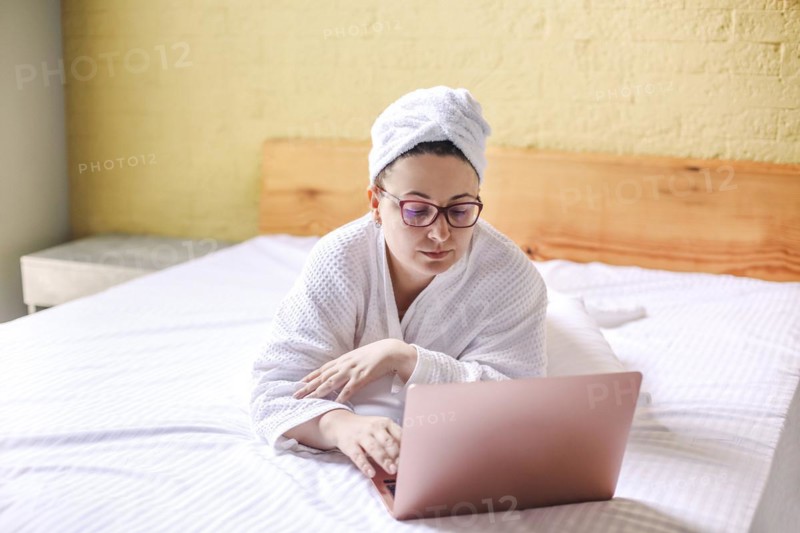 Happy woman in eyeglasses waiting and has video call on laptop lying on bed. Work from home concept