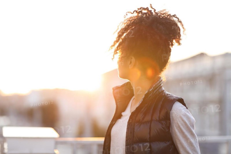 Rear view of young woman standing at rooftop in morning sunlight, african american female standing on roof and enjoying new beautiful day while looking at sun rising over house and cityscape
