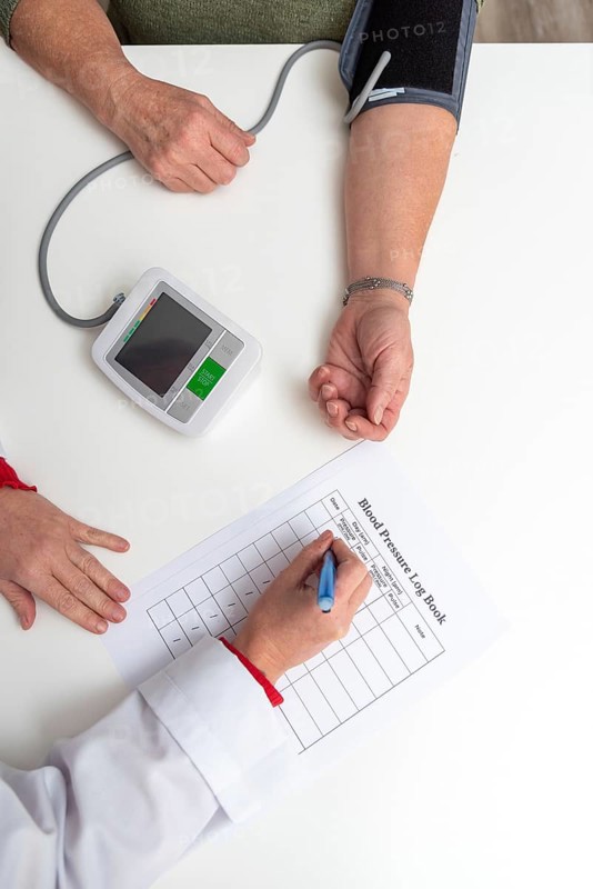 Doctor taking notes on blood pressure log book while measuring blood pressure of senior woman patient