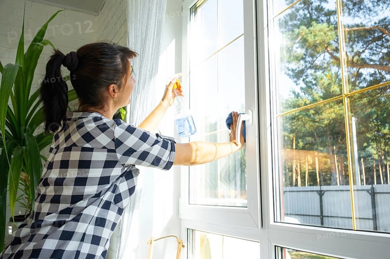 Woman manually washes the window of the house with a rag with spray cleaner and mop inside the interior with white curtains. Restoring order and cleanliness in the spring, cleaning servise