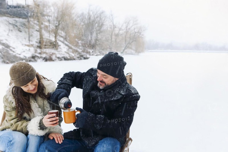 Happy lovely middle-aged couple in warm clothes drinking hot tea during romantic winter date in nature, man and woman in love sitting on frozen lake under falling snow and chatting on cold frosty day