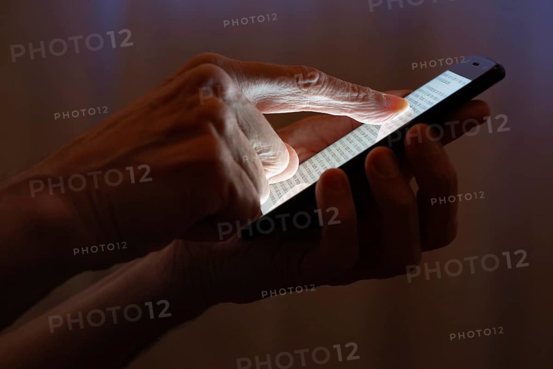 Man typing with the finger on the touch screen of a black smartphone close to the window with closed curtains at night