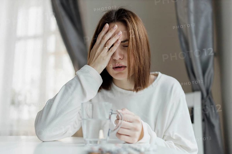 Woman holding her forehead, looking unwell, with a glass of water and curtains in the background