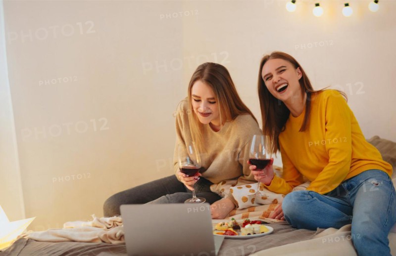 Two cute happy funny women friends having fun together, arranging home cinema evening, watching comedy on laptop, laughing out loudly, sitting on bed with cheese plate and glasses of red wine