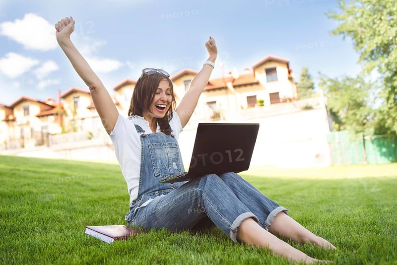 Female gets joyful emotions by talking to her friends on a conference call from notebook, she dressed in jeans overalls sitting on grass in the park